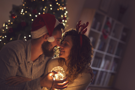 Beautiful couple in love sitting on the living room floor next to a Christmas tree, holding a jar filled with Christmas lightsの写真素材