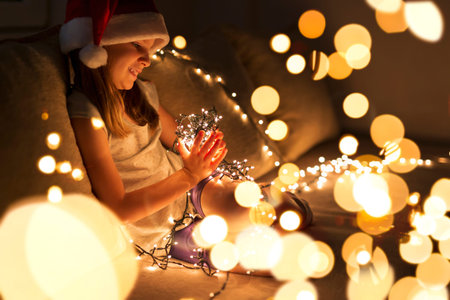 Beautiful little girl sitting on a living room couch, wearing Santa's hat and holding bunch of Christmas lights; child setting up Christmas decorations and waiting for Santaの写真素材