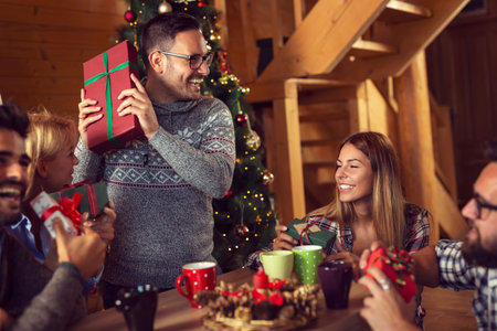 Group of young friends sitting next to a nicely decorated Christmas tree, exchanging Christmas...