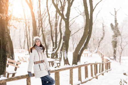 Attractive young woman leaning against a wooden fence in a park, enjoying a snowy winter day in...