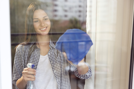 Beautiful young woman cleaning windows with a cleanser spray and a cloth; housekeeping assistant wiping windows with city reflections in the glassの写真素材