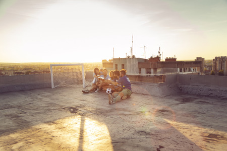 Group of young people sitting on a building rooftop, wearing jerseys, resting after a football match, drinking beer and making a toastの写真素材