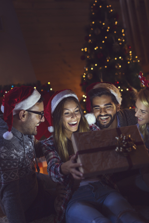 Group of friends sitting next to a fireplace and a nicely decorated Christmas tree, opening presents and having fun on Christmas Eve. Focus on the couple in the middleの写真素材
