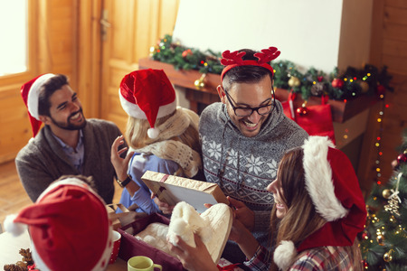 Group of young friends sitting next to a nicely decorated Christmas tree, exchanging Christmas presents and having fun. Focus on the guy in the middleの写真素材