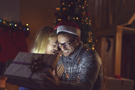Young couple in love sitting next to a fireplace and a nicely decorated Christmas tree, opening presents and having fun on Christmas Eve. Focus on the guyの写真素材
