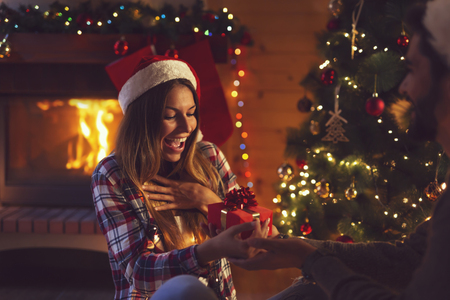 Young couple in love sitting by the fireplace and nicely decorated Christmas tree, exchanging Christmas presentsの写真素材