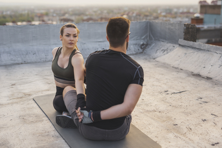 Couple in a sportswear, sitting on a yoga mat, stretching before a building rooftop terrace workoutの写真素材