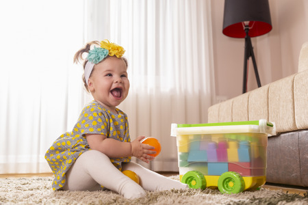Beautiful little baby girl, sitting on a carpet on the nursery floor, playing and throwing a ballの写真素材