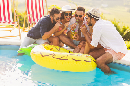 Group of friends at a poolside summer party, sitting at the edge of a swimming pool, drinking a pineapple cocktail and having funの写真素材