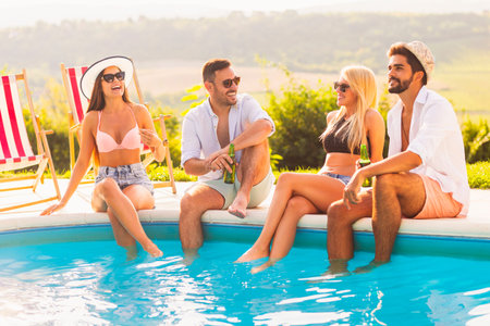 Group of friends at a poolside summer party, sitting at the edge of a swimming pool with legs in the...