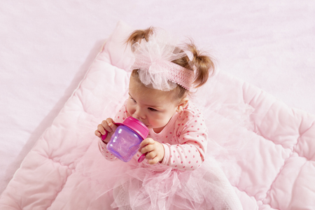 High angle view of a baby girl wearing pink tutu skirt, sitting on a pink duvet and drinking water from a bottleの写真素材