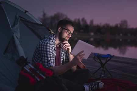 Young man camping on the lake docks, sitting on the tent entrance and reading the news on a tablet computerの写真素材