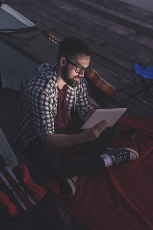 Young man camping on the lake docks, sitting on the tent entrance and working on a tablet computerの写真素材