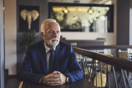 Portrait of a pensive senior businessman sitting in a modern office building restaurant, taking a coffee break and looking at the distanceの写真素材
