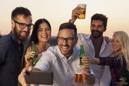 Group of friends having fun at an outdoor summertime party, dancing, drinking beer and taking selfiesの写真素材