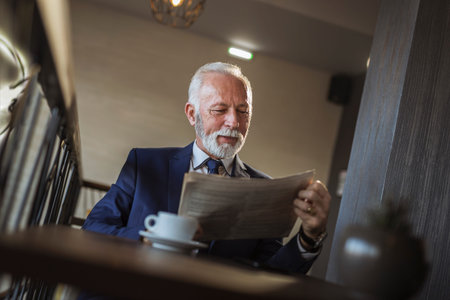 Senior businessman sitting at a restaurant table, reading newspaper and drinking coffee