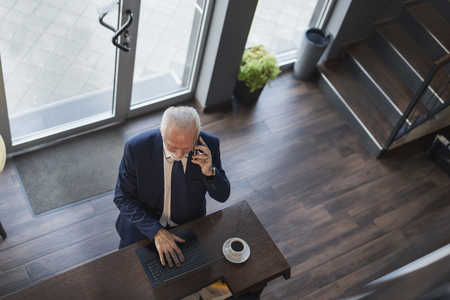 Senior businessman standing by a restaurant counter, having a phone conversation and working on a laptop computerの写真素材
