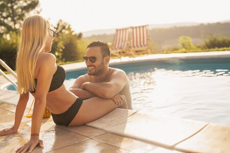 Couple in love at a poolside summer party, sitting at the edge of a swimming pool, sunbathing, drinking cocktails and having funの写真素材
