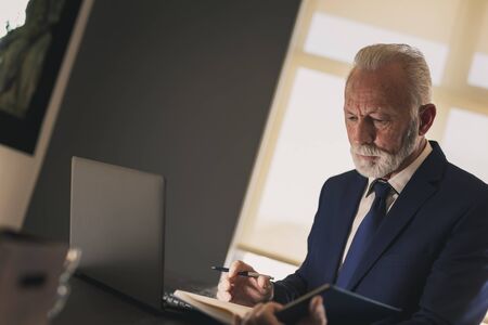 Senior businessman in a modern office working on a laptop computer and taking notes, pensive and seriousの写真素材