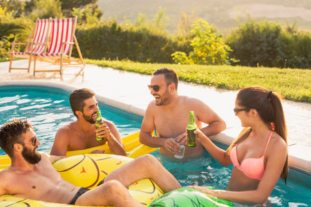 Group of friends at a poolside summer party, having fun in the swimming pool, drinking cocktails and...