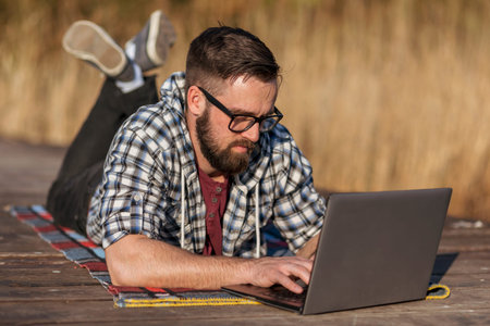 Male freelancer lying on the lake docks, working remotely while relaxing in nature