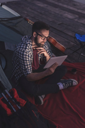 Young man camping on the lake docks, sitting on the tent entrance and working on a tablet computer