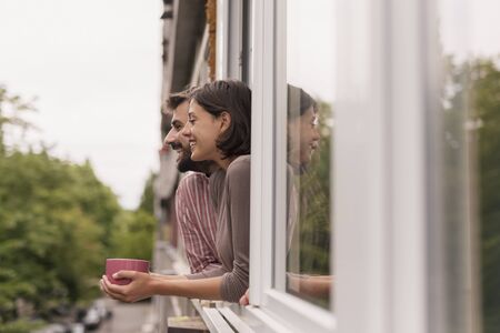 Couple in love leaning to an opened window, looking outside through it, drinking coffee and relaxing at homeの写真素材