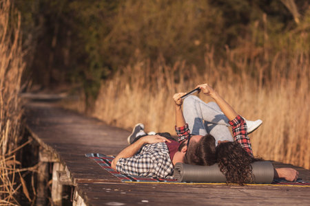 Beautiful couple in love lying on a picninc blanket at lake docks, taking a selfie and having fun on...