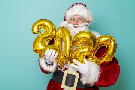 Portrait of a Santa Claus holding 2020 numbers shaped golden balloons, isolated on mint colored backgroundの写真素材