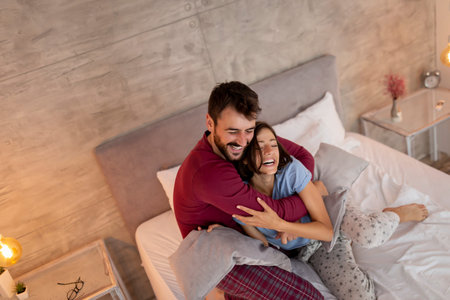 Beautiful young couple in love wearing pajamas, laughing and hugging after a pillow fight in bed