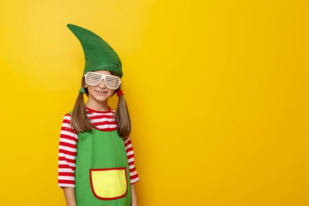 Portrait of a beautiful little girl wearing Christmas elf costume and party glasses on yellow colored background with copy spaceの写真素材