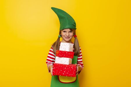 Beautiful little girl dressed as Santas helper holding a pile of nicely wrapped Christmas presents isolated on yellow colored backgroundの写真素材