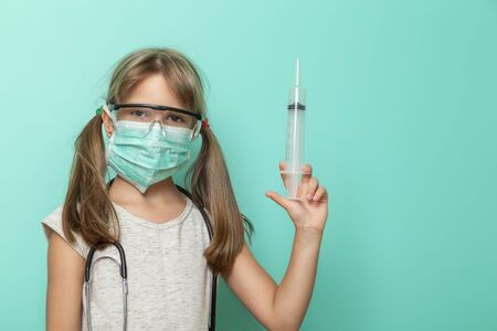 Beautiful little girl wearing surgical mask, stethoscope and protective lab glasses holding a giant syringe; child role-playing a doctor or nurseの写真素材