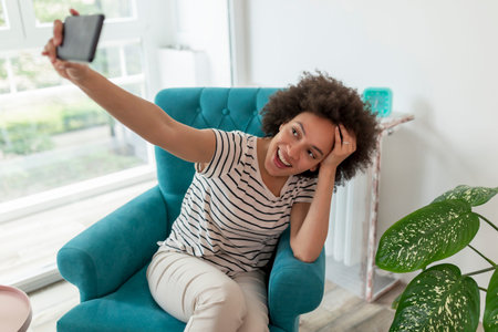 Beautiful young mixed race woman sitting in an armchair, taking a selfie with her smartphone and...