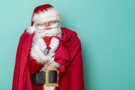 Santa Claus sick for winter holiday season, having a flu and fever, covered with soft red blanket, drinking hot tea, isolated on mint colored backgroundの写真素材