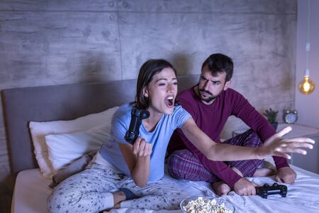 Beautiful young couple wearing pajamas, having fun at home, playing video games in bed at night, woman excited after winning the game, man disappointed after losingの写真素材