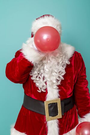Santa Claus blowing red balloon, celebrating New Year and having fun, isolated on mint colored backgroundの写真素材