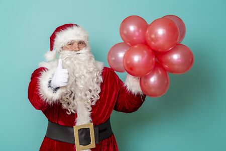 Santa Claus holding bunch of red balloons and showing thumbs up, celebrating New Year and having fun, isolated on mint colored backgroundの写真素材