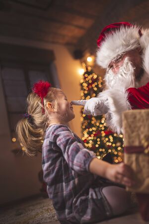 Beautiful little girl having fun at home on Christmas Eve, sitting on the carpet next to a nicely decorated Christmas tree, opening Christmas presents with Santa Clausの写真素材