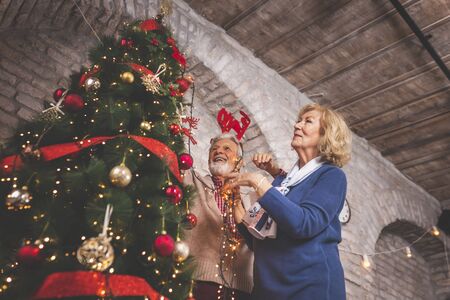 Happy senior couple standing next to a nicely decorated Christmas tree, decorating it, placing Christmas lights on it and having fun at home on Christmas dayの写真素材