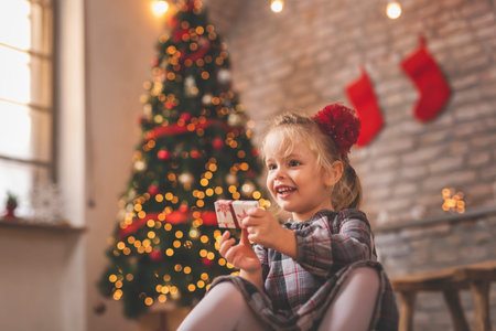 Beautiful little girl sitting by the nicely decorated Christmas tree, having fun while opening...
