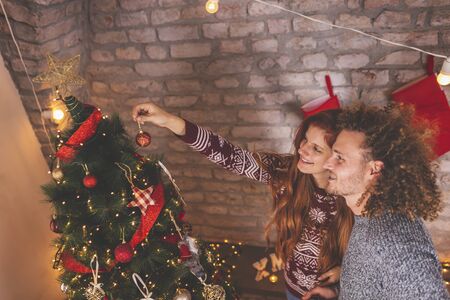 Beautiful young couple in love having fun while celebrating Christmas at home, decorating Christmas tree togetherの写真素材