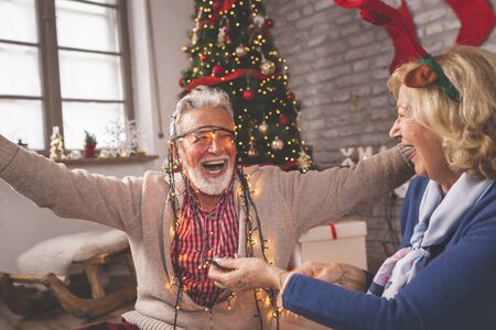 Happy senior couple sitting next to a nicely decorated Christmas tree, holding Christmas lights and having fun while decorating home on Christmas dayの写真素材