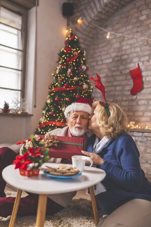Happy senior couple sitting by nicely decorated Christmas tree, celebrating Christmas at home, exchanging Christmas presents, woman kissing a manの写真素材