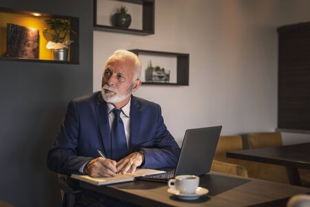 Senior businessman in a restaurant, working on a laptop computer, writing in a planner and having a cup of coffeeの写真素材