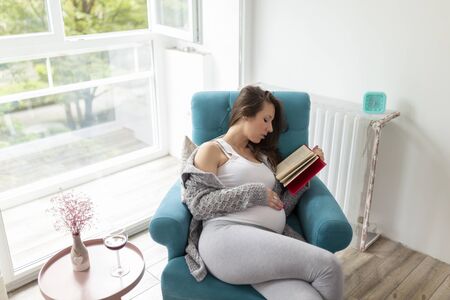 Beautiful pregnant woman sitting in an armchair by the window, reading a book and drinking blackberry wineの写真素材