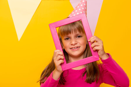 Beautiful little girl having fun celebrating her birthday, holding a pink photo frame and posing for...