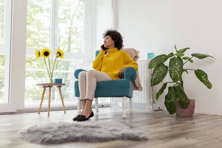 Beautiful mixed race woman sitting in an armchair, relaxing at home, having a phone conversationの写真素材