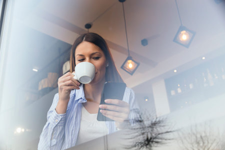 Portrait of a beautiful young woman sitting at a restaurant table, drinking coffee and typing a text...