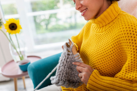 Beautiful young mixed race woman sitting in an armchair,  knitting a scarf and relaxing at home